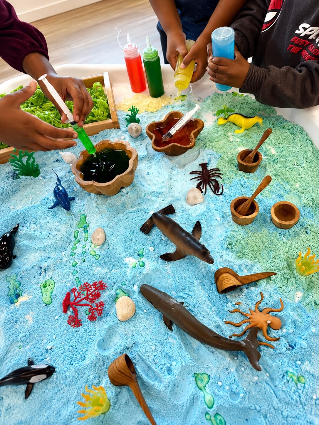 Children at a Little Hands Sensory ocean small-world tray, adding colour with syringes among toy sea creatures, shells and wooden bowls